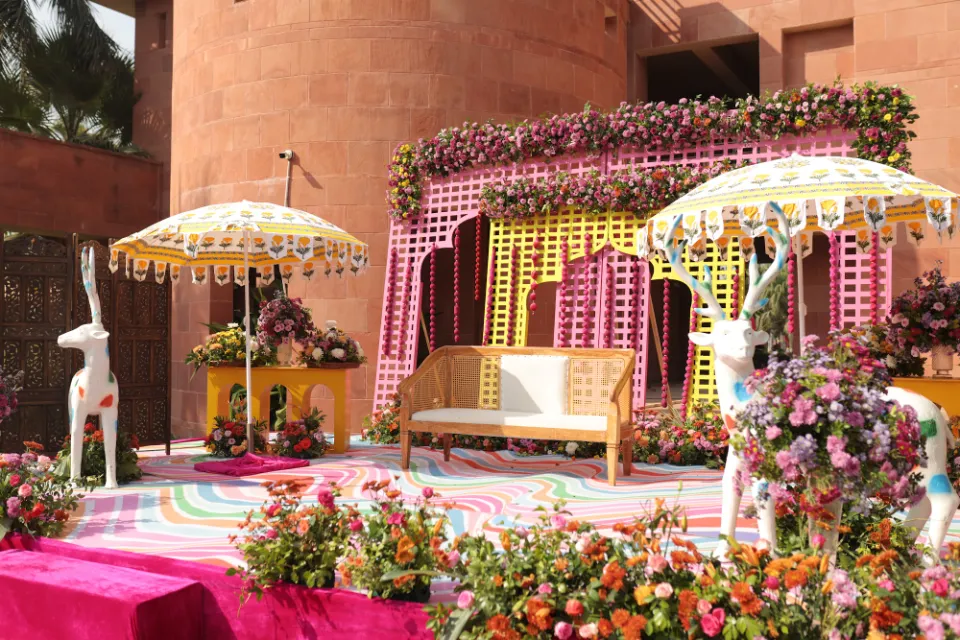 An outdoor haldi stage with a pink and yellow lattice backdrop, parasols and flowers, decorated by Bluebird Events