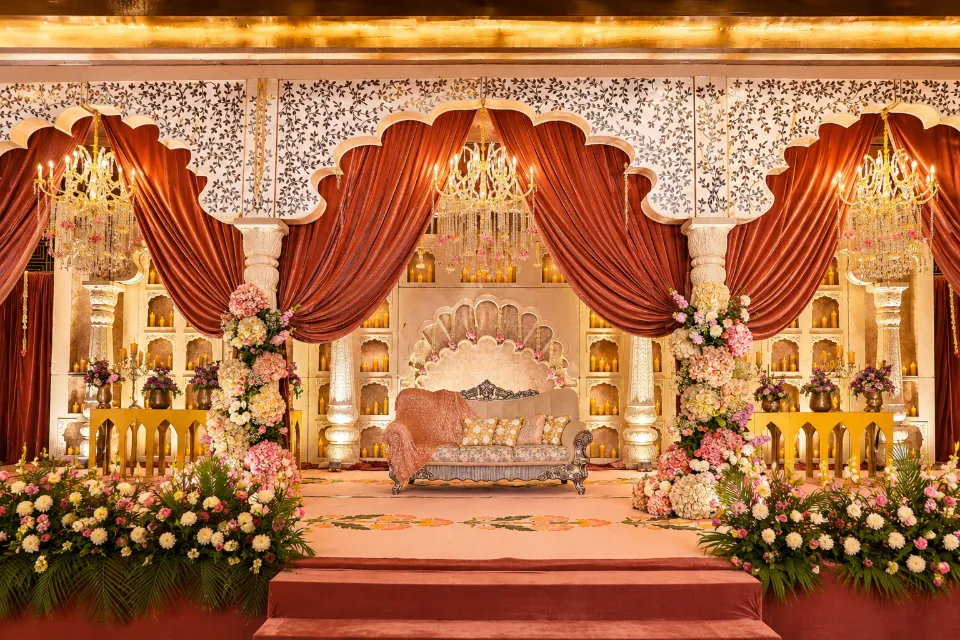 A palace-style stage in ivory and gold with velvet drapes, chandeliers and pink and white florals, decorated by Bluebird Events