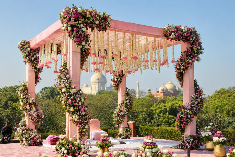 A pink floral mandap hung with marigold and rose strings, with the white dome of the Taj Mahal in the distance